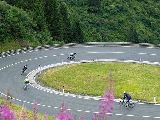 Radfahrer fahren durch eine weite Kehre mit Bergwiesen und Wald – typische Arlberg-Giro-Szene