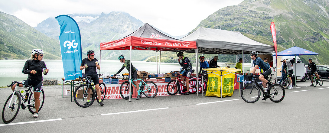 Cyclists take a break at a refreshment station with tents, banners, and snacks at the Kops reservoir with alpine mountain scenery.