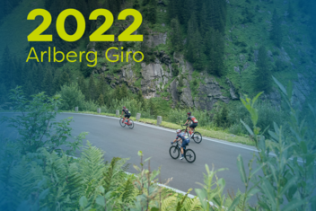Racing cyclists at the Arlberg Giro 2022 ride on wet roads through wooded areas. The athletes are wearing rain gear and helmets. ‘2022 Arlberg Giro’ is written in yellow/green at the top left of the picture.