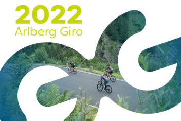 Racing cyclists at the Arlberg Giro 2022 ride on wet roads through wooded areas. The athletes are wearing rain gear and helmets. ‘2022 Arlberg Giro’ is written in yellow/green at the top left of the picture.