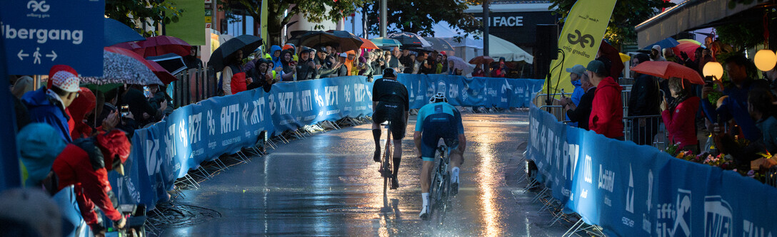 Two racing cyclists sprint along the rainy pedestrian street in the evening, spectators and advertising banners line the route. 